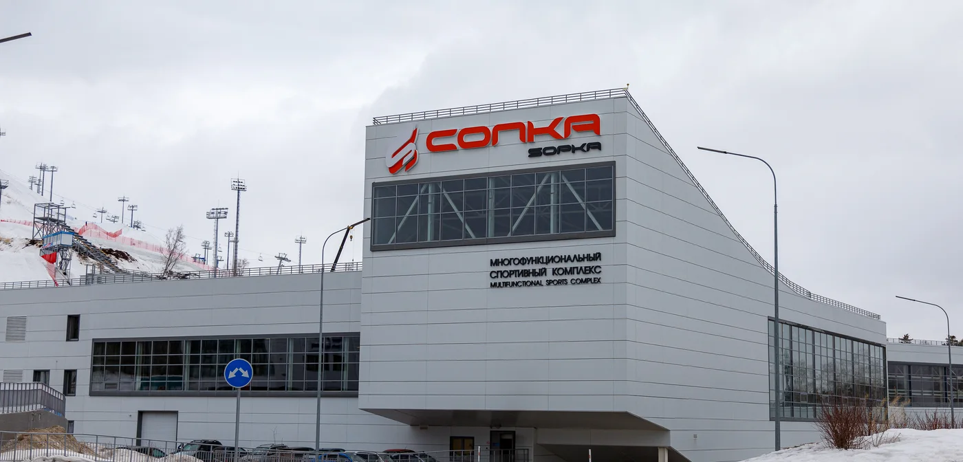 Многофункциональный спортивный комплекс «Сопка»: светлый корпус с вывеской и горнолыжная трасса с подъёмниками за ним
